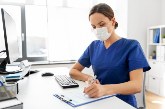 Masked clinician sitting at a desk and writing on a clipboard